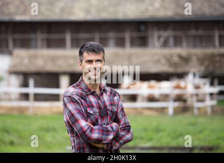 Selbstbewusster, gutaussehender Bauer mit gekreuzten Armen, der vor simmentaler Kühen und Scheunen auf der Ranch steht Stockfoto