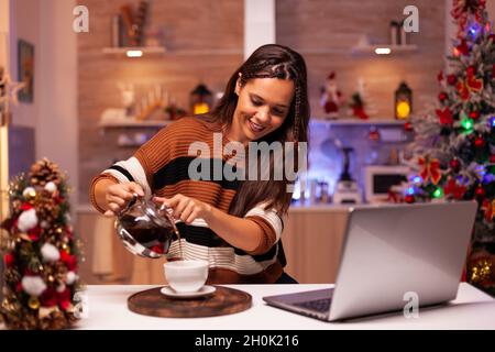 Moderne Frau füllt Tasse Tee aus dem Wasserkocher auf Videoanruf Verbindung mit Freunden zu sprechen. Fröhlicher junger Mensch, der in der saisonal eingerichteten Küche ein Notebook benutzt, um fröhliche weihnachten zu wünschen Stockfoto