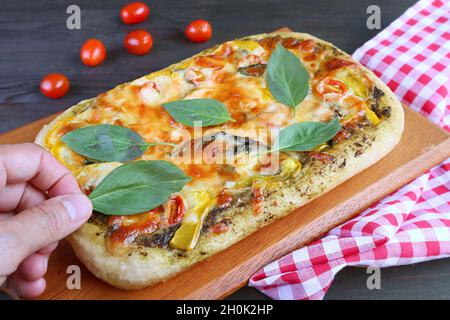 Man's Hand setzt frische Basilikumblätter auf eine köstliche frisch gebackene hausgemachte Pesto Veggie Pizza Stockfoto