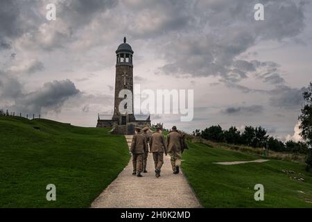 Eine Gruppe von nachwirkenden Männern in Uniformen der britischen Armee des 1. Weltkriegs, die auf einem Fußweg zu einem Kriegsdenkmal mit einem dunklen, launischen Himmel und Grasbänken gehen Stockfoto