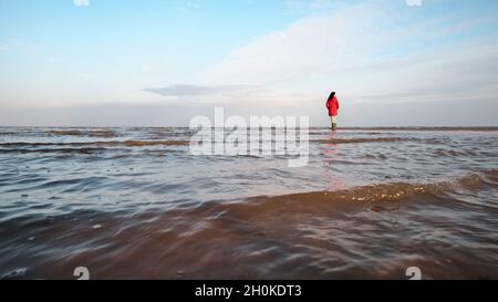 Eine Frau in einer roten Jacke schaut auf die schäumenden Wellen. Windiger Herbsttag am Ufer des Ilmensees. Stockfoto