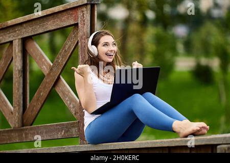 Eine fröhliche Frau mit kabellosen Kopfhörern macht im Freien Telearbeit mit einem Laptop. Stockfoto