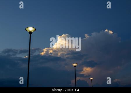 Ein Detail von drei Straßenlaternen mit dem Licht an und bewölktem und dunklem Himmel im Hintergrund. Stockfoto