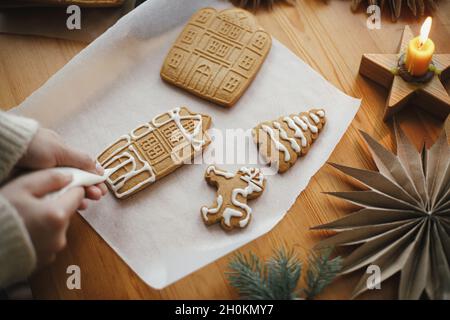 Hände schmücken weihnachts-Lebkuchenkekse mit Glasur auf Holztisch mit Kerze und Ornamenten. Nahaufnahme. Herstellung Lebkuchenhaus mit Zuckerguss. Stockfoto
