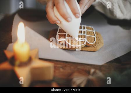 Hände schmücken weihnachts-Lebkuchenkekse mit Glasur auf Holztisch mit Kerze und Ornamenten. Nahaufnahme der Herstellung von Lebkuchenhaus mit Frostin Stockfoto