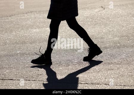 Silhouette und Schatten des Mädchens, das die Straße entlang geht. Weibliche Beine auf dem Bürgersteig, eilende Frau im Herbstmantel, Konzept der Einsamkeit Stockfoto