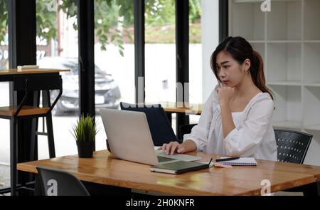 Fokussierte asiatische Geschäftsfrau, die im Büro online studiert und dabei einen Laptop betrachtet, seriöse Angestellte oder Studenten, die sich das Webinar ansehen und Informationen schreiben Stockfoto