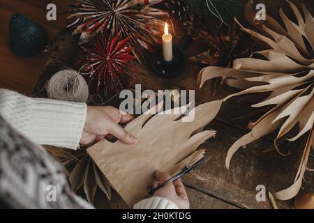 Stylische Weihnachtssterne. Hände schneiden Stern aus Bastelpapier Tasche auf Hintergrund von handgefertigten schweden Sterne, Faden, Kerze, Schere auf rustikalen woo Stockfoto