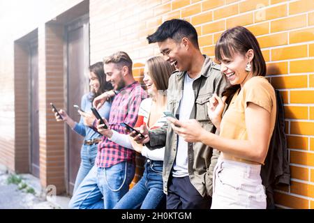 Multikulturelle Freunde lachen mit dem Smartphone an der Wand auf dem Universitätscampus - Junge Menschen süchtig nach mobilen Smartphones - Technologie conce Stockfoto