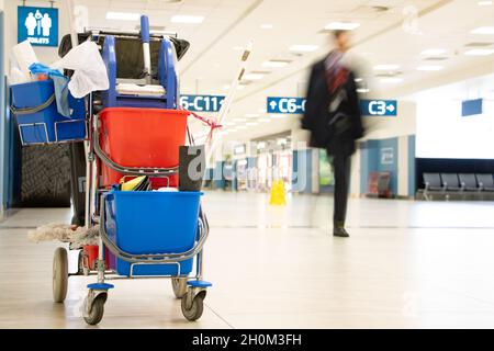 Ein Servicewagen mit Reinigungszubehör in einem leeren Flughafenterminal mit verschwommenen Menschen auf einem Hintergrund. Stockfoto