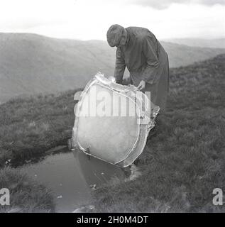 Die 1950er Jahre, historisch, winterfest und ein Mann in einem Regenmantel und Stoffkappe hoch oben auf einem nassen kalten Hügel im Lake District, Cumbria, England, Großbritannien, der einen festen Eisblock vom Boden hebt, um einen Pool oder Brunnen mit Wasser darunter zu enthüllen. Stockfoto