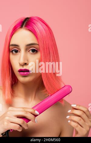 Junge Frau mit gefärbtem Haar mit Haarglätter isoliert auf rosa Stockfoto