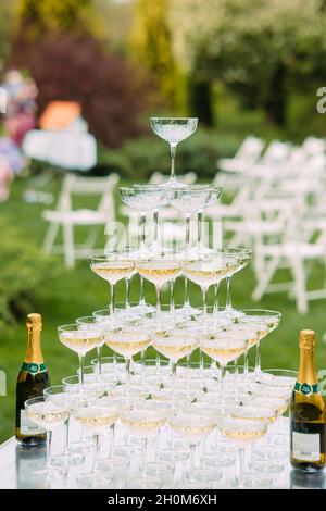 Zwei Flaschen Champagner und Weingläser mit Pyramide stehen auf dem mit weißer Tischdecke überzogenen Tisch beim Hochzeitsbankett vor dem Hintergrund von Rasen und Ga Stockfoto