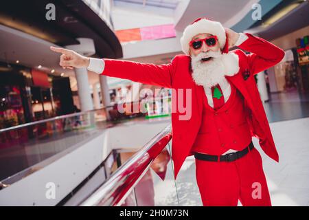 Foto von Mode trendy beeindruckt santa claus tragen dunkle Brillen zeigen Finger weit weg lächelnd drinnen Boutique Store Stockfoto