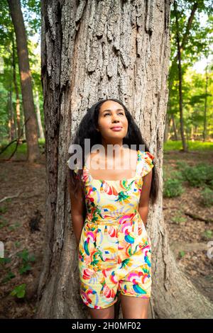 Wunderschönes Porträt einer glücklichen, gemischten afroamerikanischen Frau mit einem gelben Blumenkleid, das sich an einen großen, mit rauen Rinden bedeckten Baumstamm lehnt Stockfoto