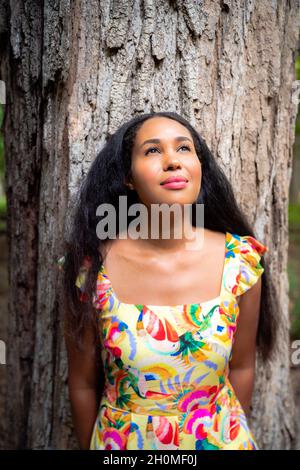 Wunderschönes Porträt einer glücklichen, gemischten afroamerikanischen Frau mit einem gelben Blumenkleid, das sich an einen großen, mit rauen Rinden bedeckten Baumstamm lehnt Stockfoto