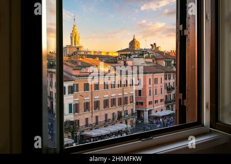 Blick durch ein offenes Fenster über der Piazza Navona bei Sonnenuntergang auf die farbenfrohen Cafés, Türme und Kirchen entlang der Skyline. Stockfoto