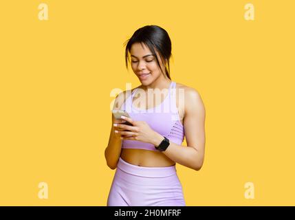Fit afroamerikanische Frau in Sportuniform mit Smartphone, Chat mit Personal Trainer, gelber Hintergrund Stockfoto