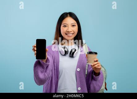 Aufgeregt asiatische Studentin zeigt Smartphone mit leerem Bildschirm, posiert mit Rucksack und Kaffee, mockup Stockfoto
