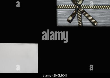 Unschärfe-Effekt: Ein Geschenk mit goldener Schleife und leerer weißer Karte auf dunkelschwarzem Hintergrund. Horizontal mit Kopierbereich. Graue männliche Geschenkbox für ihn, Mann, Männer, Vater Stockfoto