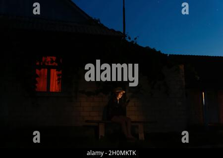 Unschärfe-Effekte für Frauen mithilfe von Smartphones, die nachts auf der Bank in der Nähe des Hauses sitzen. Außenportrait von Millennial, das nachts ihr Mobiltelefon benutzt. Orangefarbenes Licht in w Stockfoto