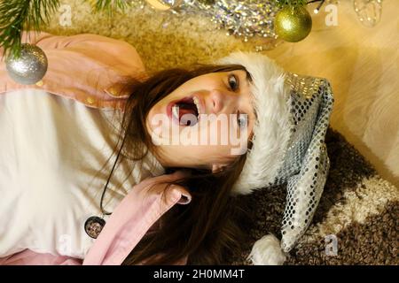 Porträt einer verrückten, überraschten jungen Brunet Frau 20-25 Jahre in weißem weihnachtsmann Hut und rosa Hemd. Auf dem Teppich unter dem Weihnachtsbaum auf dem Hintergrund liegend. Stockfoto