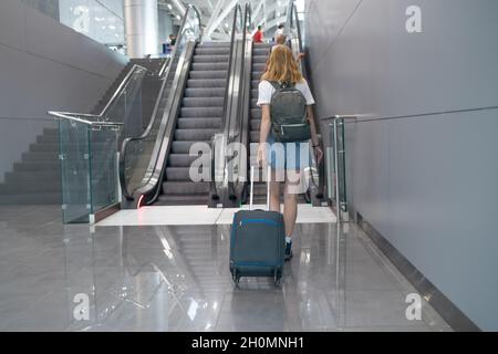 Blonde, haarige Touristin mit Rucksack und Koffer fährt mit der Rolltreppe zum Check-in an einem internationalen Flughafen. Reisekonzept Stockfoto