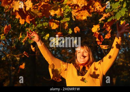 Hallo Herbst. Fröhliche blonde elegante 40-jährige Frau in gelbem Pullover, Schal draußen im Herbstpark genießen Herbst und Blätter werfen. Viele fl Stockfoto