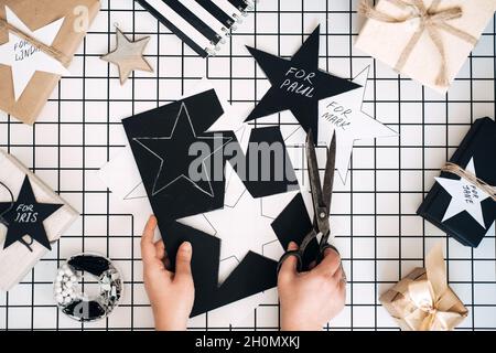 Personalisierte Geschenke. Modernes Schwarz-Weiß-Flachlappen mit Geschenkverpackung für Weihnachten. Weibliche Hand mit Schere, eingewickelte Bastelkisten mit Namen Stockfoto