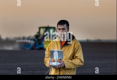 Attraktiver Landwirt, der Drohne über Ackerland steuert. Traktor arbeitet im Hintergrund. Hochtechnologische Innovationen zur Steigerung der Produktivität in der Landwirtschaft Stockfoto