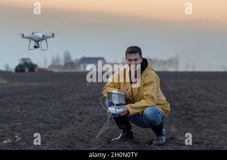 Attraktiver Landwirt, der Drohne über Ackerland steuert. Traktor arbeitet im Hintergrund. Hochtechnologische Innovationen zur Steigerung der Produktivität in der Landwirtschaft Stockfoto