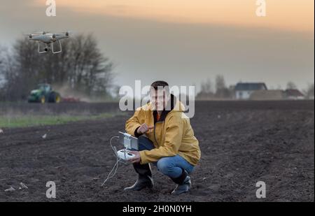 Attraktiver Landwirt, der Drohne über Ackerland steuert. Traktor arbeitet im Hintergrund. Hochtechnologische Innovationen zur Steigerung der Produktivität in der Landwirtschaft Stockfoto