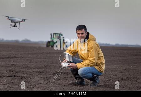 Attraktiver Landwirt, der Drohne über Ackerland steuert. Traktor arbeitet im Hintergrund. Hochtechnologische Innovationen zur Steigerung der Produktivität in der Landwirtschaft Stockfoto