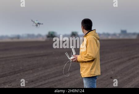 Attraktiver Landwirt, der Drohne über Ackerland steuert. Traktoren arbeiten im Hintergrund. Hightech-Innovationen zur Steigerung der Produktivität in agricult Stockfoto