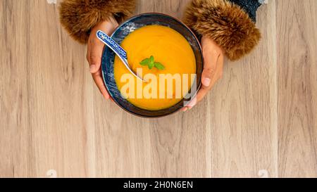 Hände halten Kürbissuppe auf dem Tisch. Weibliche Hände halten eine Schüssel Kürbissuppe mit Minzblatt. Essen der Saison. Stockfoto
