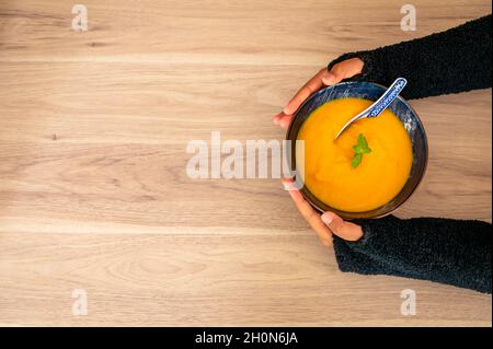 Hände halten Kürbissuppe auf dem Tisch. Weibliche Hände halten eine Schüssel Kürbissuppe mit Minzblatt. Essen der Saison. Stockfoto