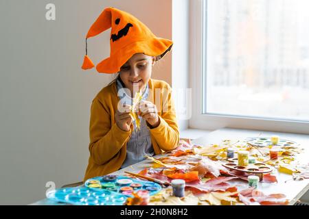 Kaukasisches Mädchen in einem Hexenkostüm sitzt zu Hause am Tisch und bereitet Kunsthandwerk für den Halloween-Urlaub vor. DIY und Dekor im Zimmer gemacht Stockfoto