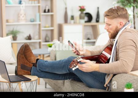 Mann spielt akustische Gitarre und schaut auf das Smartphone Stockfoto