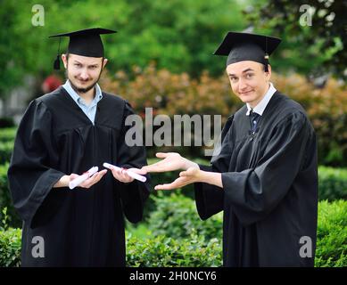 Zwei junge Hochschulabsolventen in Gewändern und quadratischen Hüten freuen sich über ein Diplom. Stockfoto