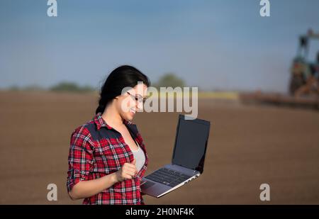 Junge glückliche Farmerin Mädchen auf dem Feld mit Laptop im Frühjahr Zeit stehen. Traktor arbeitet im Hintergrund Stockfoto