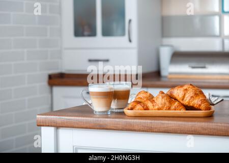 Croissants und zwei Kaffee mit Milch zur Frühstückszeit auf braunem Küchentisch. Ein Paar Cappuccino in durchsichtigen Tassen und französische Croissants an Bord Stockfoto