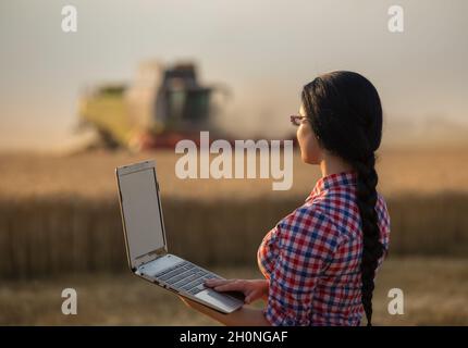 Junge Bäuerin mit Laptop, die bei Sonnenuntergang vor dem Mähdrescher auf dem Feld steht Stockfoto