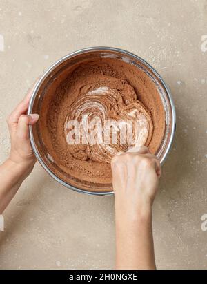 Prozess der Herstellung von Teig für Schokoladenkuchen. Mischen von Kakaopulver in Metallschüssel mit Teig auf Küchentisch, Draufsicht Stockfoto