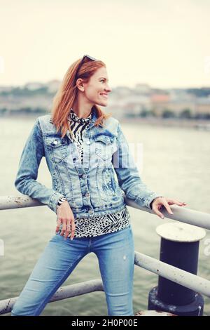 Glückliche positive Rotschopf kaukasische Frau in Denim Jacke und blaue Jeans posiert am Flussufer Stockfoto