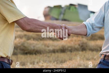 Zwei Bauern schüttelten sich während der Ernte die Hände auf das Weizenfeld. Mähdrescher arbeiten im Hintergrund Stockfoto