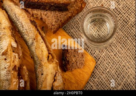 Ein kleiner gebratener Fisch auf einem Holzbrett, Brotscheiben und ein Glas Wodka, Nahaufnahme, selektiver Fokus Stockfoto