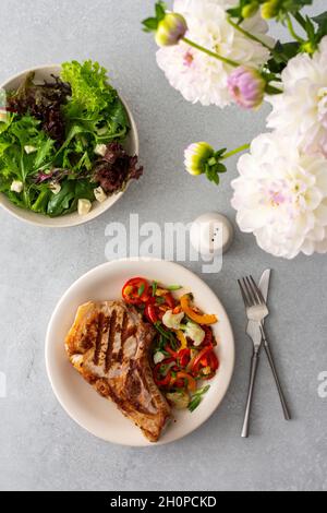 Köstliches herzhaftes Abendessen auf Tellern auf dem Tisch und ein Bouquet von schönen Herbstblumen, gebratenes Schweinefleisch-Entrecote auf dem Knochen mit geschmortem Gemüse Stockfoto