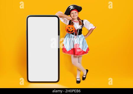 Halloween. Porträt eines Kindes im Karnevalskostüm eines Piraten im Studio auf gelbem Hintergrund. Ein kleines Mädchen steht neben einem großen Smartphone Stockfoto