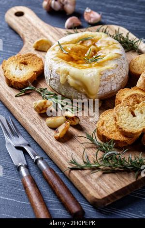 Im Ofen gebackener französischer Camembert-Käse mit Rosmarinzweigen, Knoblauchzehen und Lorbeerblättern, serviert mit Croutons auf einem Holzbrett auf einem dunklen Holzbrett Stockfoto