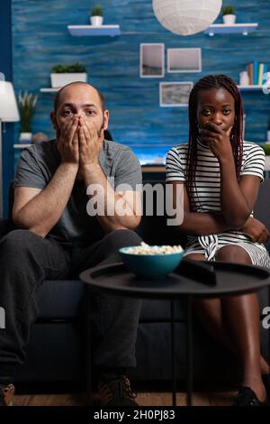 POV des interracial Paares, das schockiert ist, als es im Wohnzimmer einen Theaterfilm im Fernsehen ansieht. Gemischte Rennpartner mit den Händen über dem Mund und Blick auf Kamera und Fernsehen. Multiethnische Liebhaber Stockfoto